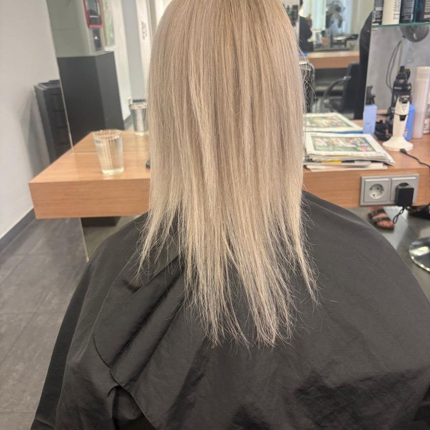 Eine Frau mit langen blonden Haaren sitzt in einem Friseurstuhl, mit einem Schreibtisch und Zeitschriften hinter ihr.