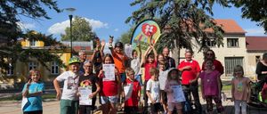 Eine Gruppe von Kindern und Erwachsenen steht auf einer Plattform und hält Urkunden in der Hand. Sie befinden sich in einer sonnigen Außenumgebung mit Gebäuden und Bäumen im Hintergrund.