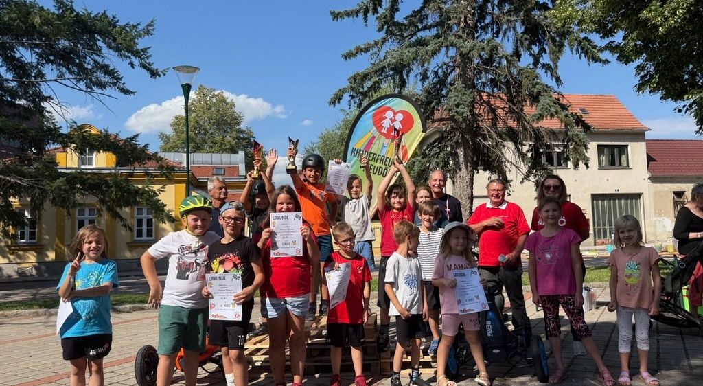 Eine Gruppe von Kindern und Erwachsenen steht auf einer Plattform und hält Urkunden in der Hand. Sie befinden sich in einer sonnigen Außenumgebung mit Gebäuden und Bäumen im Hintergrund.