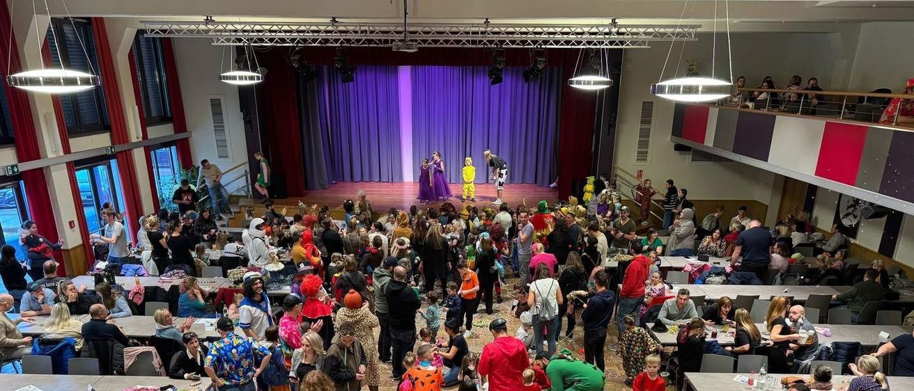 Eine große Menschenmenge, einschließlich Kinder in Kostümen, ist in einem Saal versammelt. Der Saal hat Tische mit Gegenständen und eine Bühne mit Darstellern. Konfetti ist auf dem Boden verstreut.
