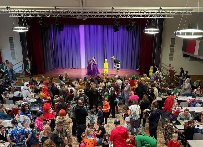 Eine große Menschenmenge, einschließlich Kinder in Kostümen, ist in einem Saal versammelt. Der Saal hat Tische mit Gegenständen und eine Bühne mit Darstellern. Konfetti ist auf dem Boden verstreut.