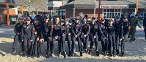 Eine Gruppe junger Menschen in schwarzen Uniformen posiert für ein Foto auf einem Kiesplatz vor einem Schulgebäude.