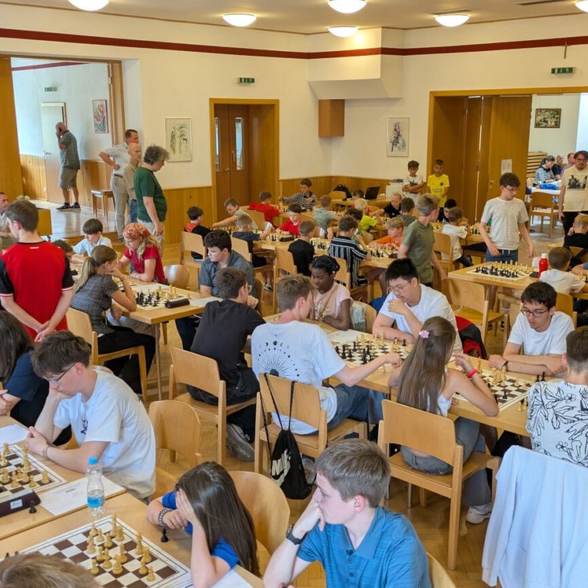 A large group of young people are playing chess in a room with many tables. The room has wooden floors and white walls.