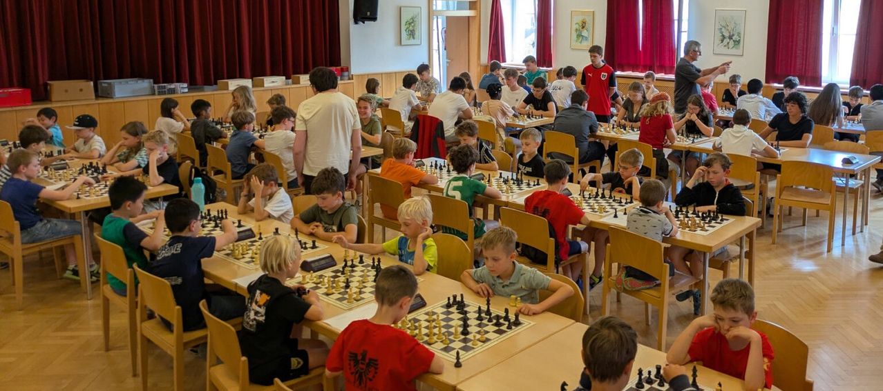 A group of children and adults play chess in a room with wooden chairs, tables, and a stage with a speaker.
