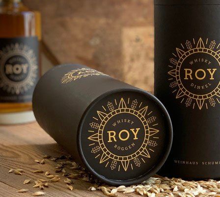 Ein Holztisch hält zwei Flaschen Roy Whisky und zwei zylindrische Dosen mit goldenen Logos und dem Wort Roy darauf. Die Dosen sind aufrecht und seitlich platziert, mit Körnern drumherum.