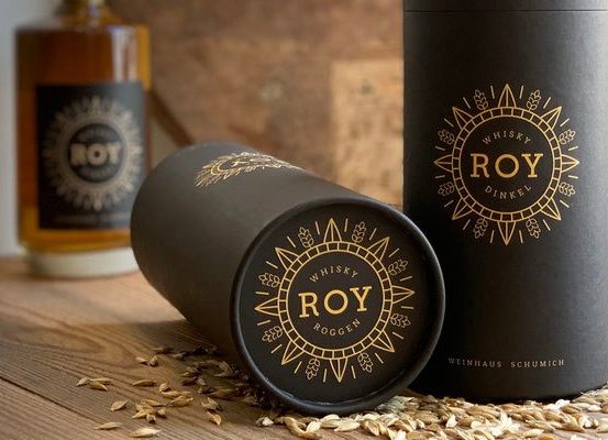 Ein Holztisch hält zwei Flaschen Roy Whisky und zwei zylindrische Dosen mit goldenen Logos und dem Wort Roy darauf. Die Dosen sind aufrecht und seitlich platziert, mit Körnern drumherum.