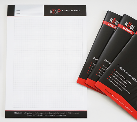 Ein Satz Geschäftsmaterialien besteht aus einem gelogoten Notizbuch, Broschüren und einem Karabiner. Das Notizbuch hat ein Rastermuster und ist neben drei Broschüren platziert. Der Karabiner befindet sich auf der linken Seite des Bildes.