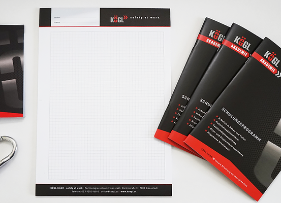 Ein Satz Geschäftsmaterialien besteht aus einem gelogoten Notizbuch, Broschüren und einem Karabiner. Das Notizbuch hat ein Rastermuster und ist neben drei Broschüren platziert. Der Karabiner befindet sich auf der linken Seite des Bildes.