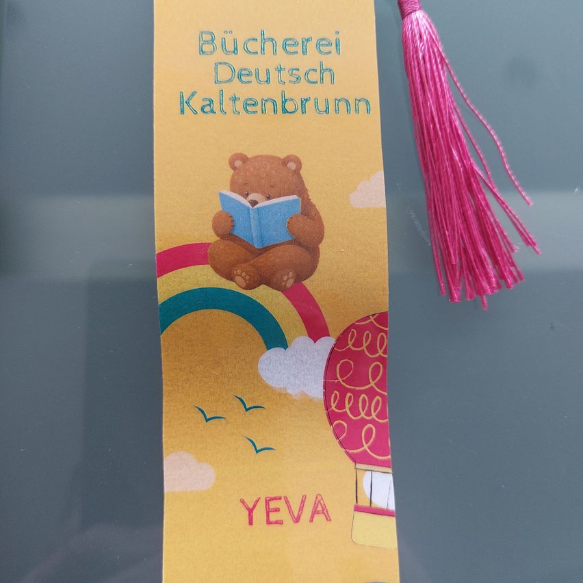 Ein gelber Lesezeichen mit einem braunen Bären, der ein Buch liest, einem Regenbogen und einem Heißluftballon. Der Text lautet 'Bucherei Deutsch Kaltenbrunn' und 'YEVA'.