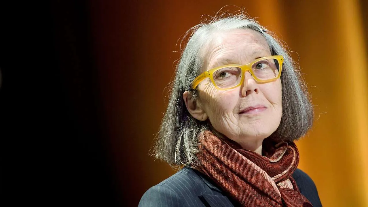 Eine ältere Frau mit grauem Haar, die eine Brille und einen Schal trägt, schaut direkt in die Kamera.