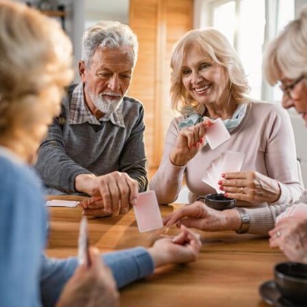 Eine Gruppe älterer Personen sitzt um einen Holztisch herum und spielt Karten. Sie scheinen es zu genießen, lächeln und lachen.