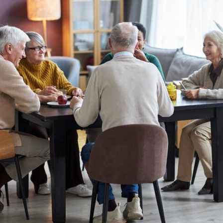 Eine Gruppe älterer Erwachsener versammelt sich um einen Tisch und führt ein Gespräch. Sie lächeln und scheinen die Gesellschaft des anderen zu genießen.