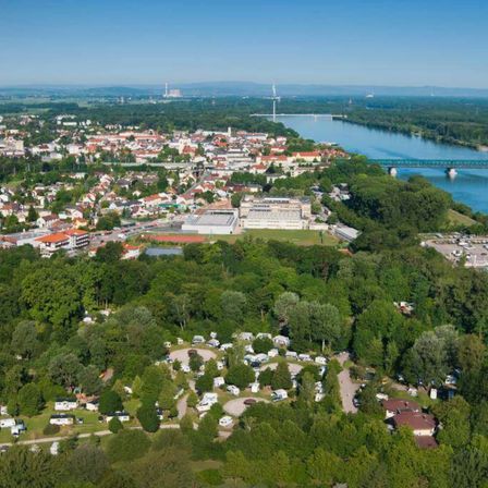 Luftaufnahme einer Stadt mit vielen Gebäuden, Bäumen und einem Fluss. Eine Brücke überquert den Fluss in der Ferne.