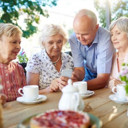 Vier ältere Erwachsene sitzen um einen Tisch, lächeln und schauen auf ein Handy. Sie haben Tassen, Untertassen und eine Teekanne auf dem Tisch. Vor ihnen steht ein Dessert.