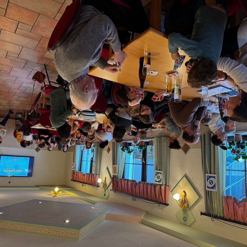 Eine Gruppe von Menschen ist in einem Raum versammelt und schaut nach oben, möglicherweise spielen sie ein Spiel. Es gibt Stühle und einen Tisch mit Flaschen und Gläsern. Eine Anzeigetafel mit Text ist an der Wand montiert.