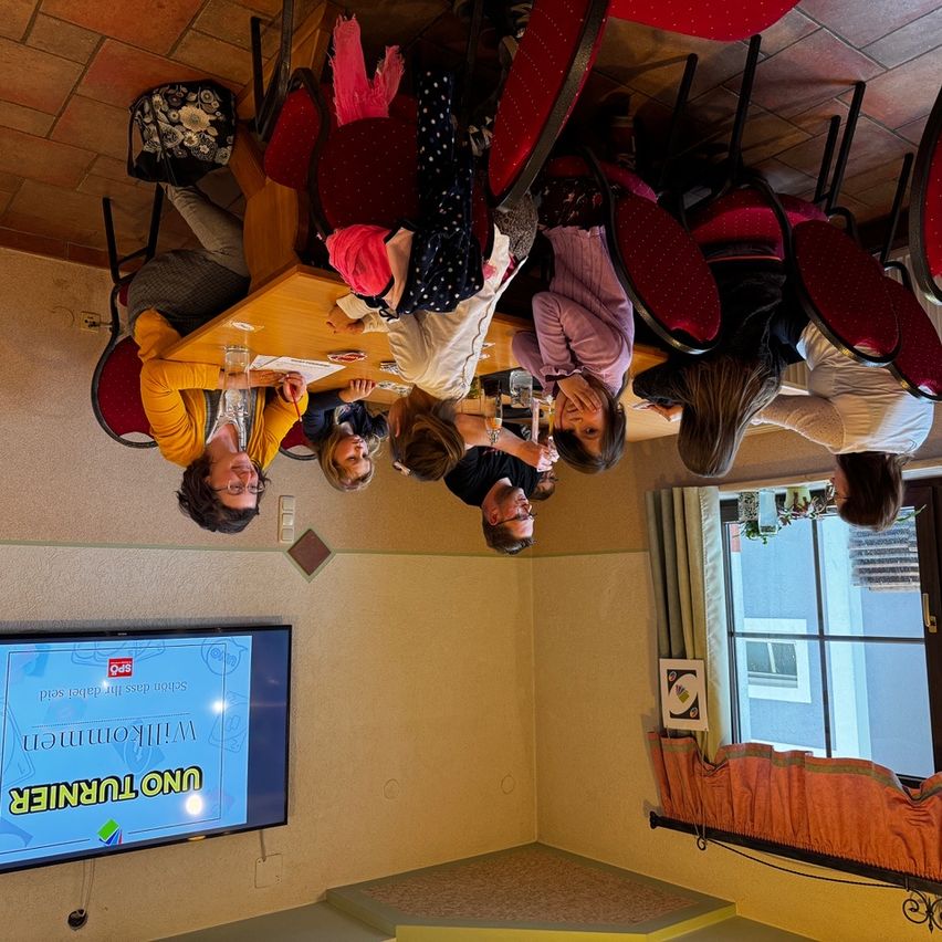 Eine Gruppe von Menschen sitzt um einen Tisch herum und schaut auf einen Bildschirm, der 'Mathematik-Challenge' anzeigt. Sie scheinen an einer mathematischen Herausforderung teilzunehmen.