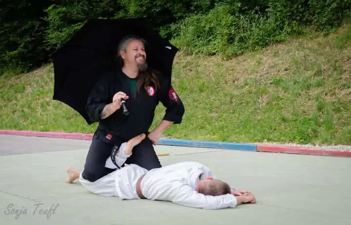 Ein Kampfkünstler hält einen Regenschirm, während er auf dem Boden kniet und einen anderen Menschen umarmt. Sie befinden sich auf einem Mattenboden mit Grünflächen und Gehwegen im Hintergrund.