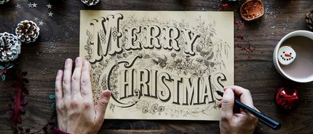 Eine Person hält eine Weihnachtskarte mit handgezeichneten Buchstaben und umgeben von Weihnachtsdekoration.