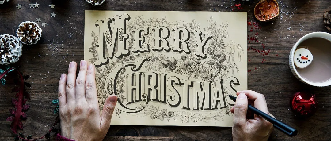 Eine Person hält eine Weihnachtskarte mit handgezeichneten Buchstaben und umgeben von Weihnachtsdekoration.