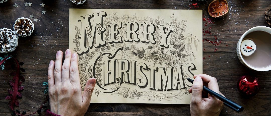 Eine Person hält eine Weihnachtskarte mit handgezeichneten Buchstaben und umgeben von Weihnachtsdekoration.