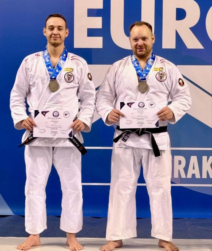 Zwei Männer in weißen Kampfsportuniformen stehen nebeneinander mit Medaillen um den Hals. Sie halten Urkunden in den Händen und lächeln für ein Foto.