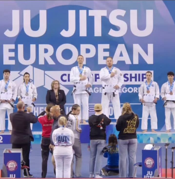 Mehrere Athleten stehen auf dem Podium bei der Ju-Jitsu-Europameisterschaft, tragen Medaillen und Uniformen. Sie werden von Offiziellen und Zuschauern begleitet, vor einem Hintergrund mit dem Namen der Veranstaltung.