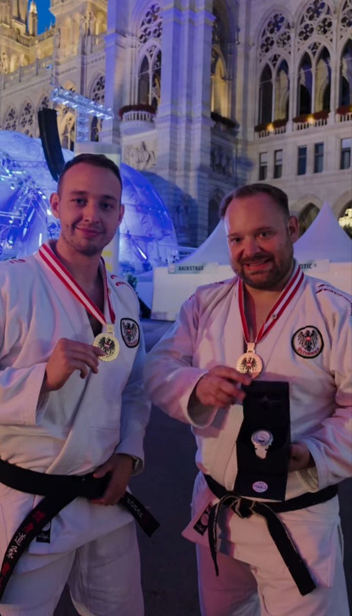 Zwei Männer in weißen Karate-Uniformen und Medaillen um den Hals lächeln für ein Foto bei einer Veranstaltung, mit einem weißen Zelt und einem Gebäude im Hintergrund.