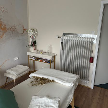 Ein kleiner Raum mit einem Bett, einem Heizkörper und einem Tisch mit Blumenarrangement. Das Bett ist mit weißen Handtüchern bedeckt.