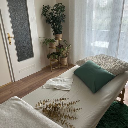 Ein Massagetisch mit einem weißen Laken, einem grünen Kissen und einem Handtuch steht in einem Raum mit Holzboden. Topfpflanzen stehen auf einem goldenen Ständer neben einer weißen Tür.