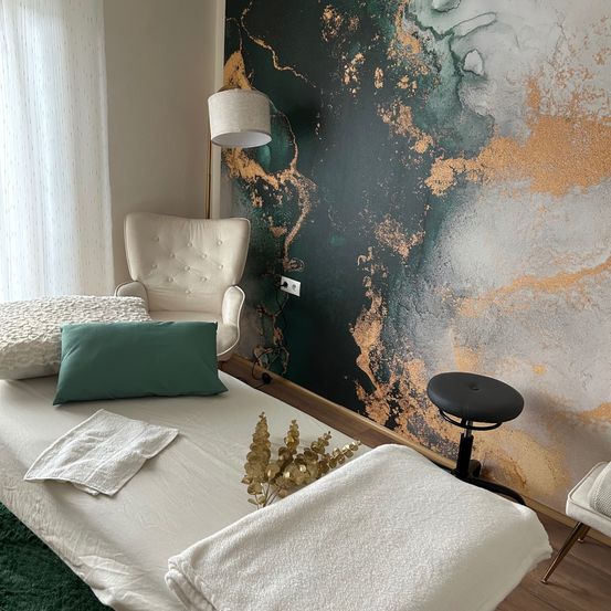 Ein kleiner Raum mit einem Bett, einem Stuhl und einer dekorativen Wand. Das Bett hat weiße Laken, Kissen und Handtücher. Eine Lampe steht an der Seite und ein Hocker steht in der Nähe der Wand.