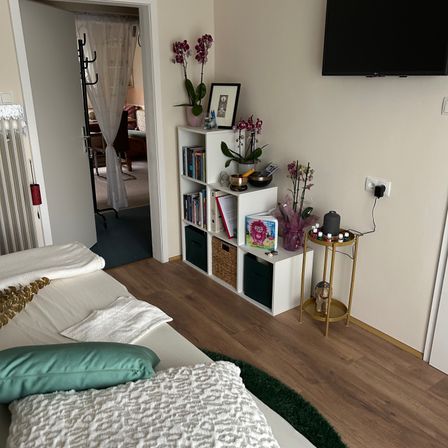 Ein Schlafzimmer mit einem Bett, einem Regal mit Büchern, Blumen und einem Fernseher an der Wand.
