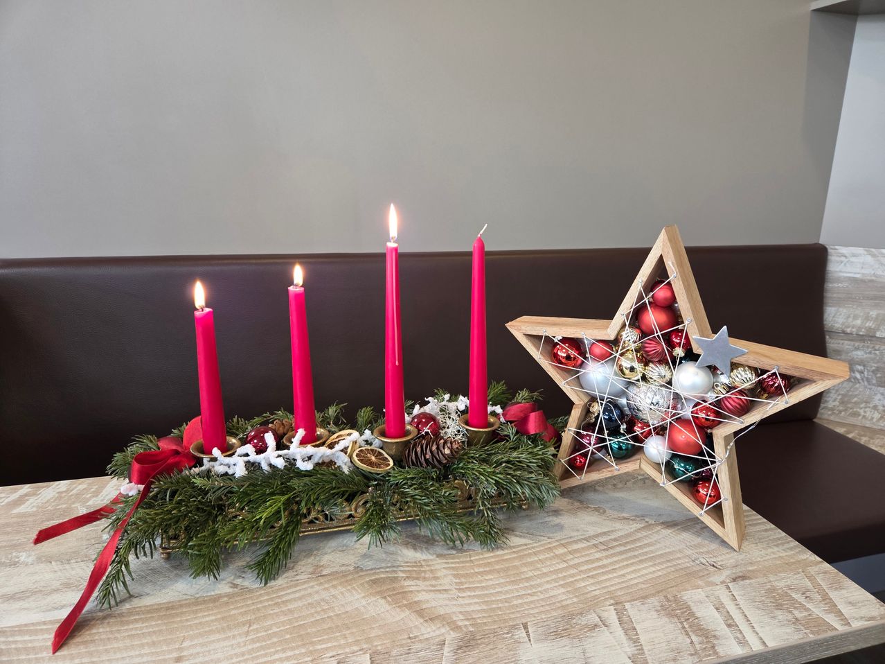 Vier rote Kerzen sind auf einem Holztisch angeordnet, begleitet von einem sternförmigen Ornament, das mit verschiedenen bunten Ornamenten gefüllt ist.