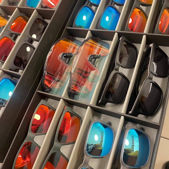 Ein Vitrinendisplay mit verschiedenen Paaren von Sonnenbrillen in unterschiedlichen Farben, einige mit reflektierenden Gläsern. Die Gläser sind ordentlich in Reihen angeordnet und präsentieren die Vielfalt der verfügbaren Farbtöne.