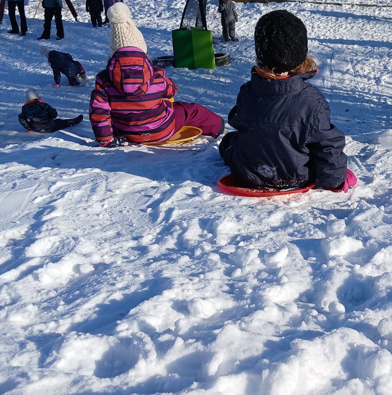 Mehrere Kinder sitzen auf Schlitten auf einer verschneiten Oberfläche. Ein Kind liegt auf dem Boden, während andere dahinter stehen. Im Hintergrund befinden sich auch Mülleimer und andere Objekte.