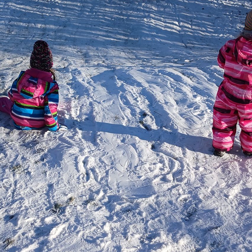 Zwei Kinder in Winterkleidung sitzen in einer verschneiten Landschaft. Eines trägt einen rosa Schneeanzug, das andere ein rosa und mehrfarbiges Jackett. Beide sind von Reifenspuren im Schnee umgeben.