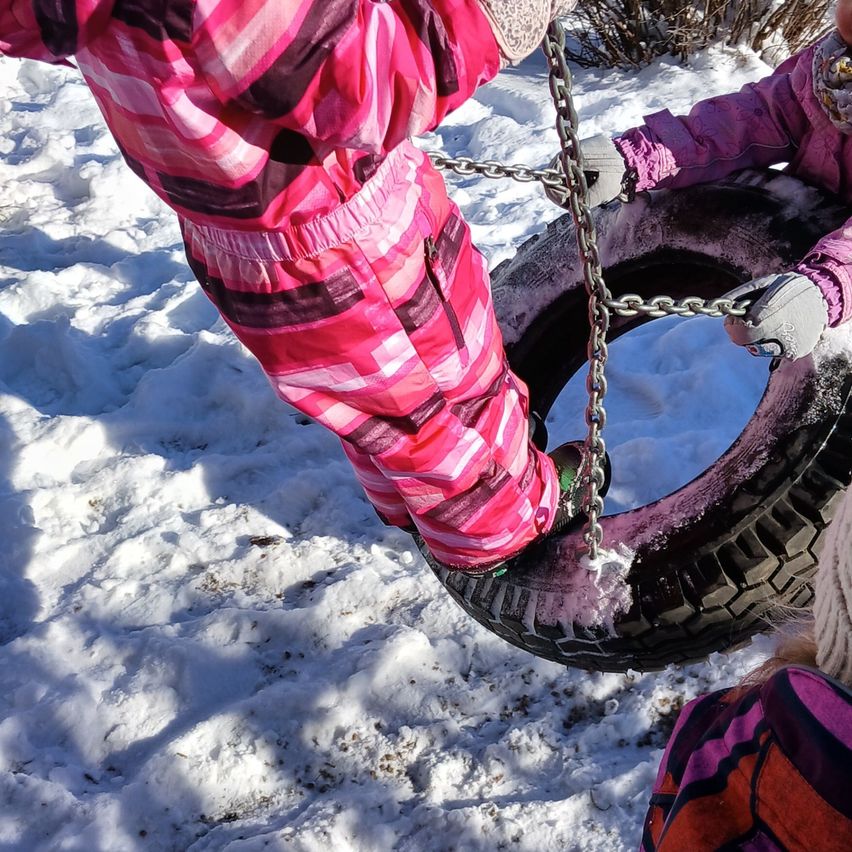Zwei Kinder in rosa und schwarzen Winterklamotten spielen auf einer Reifenschaukel im Schnee.