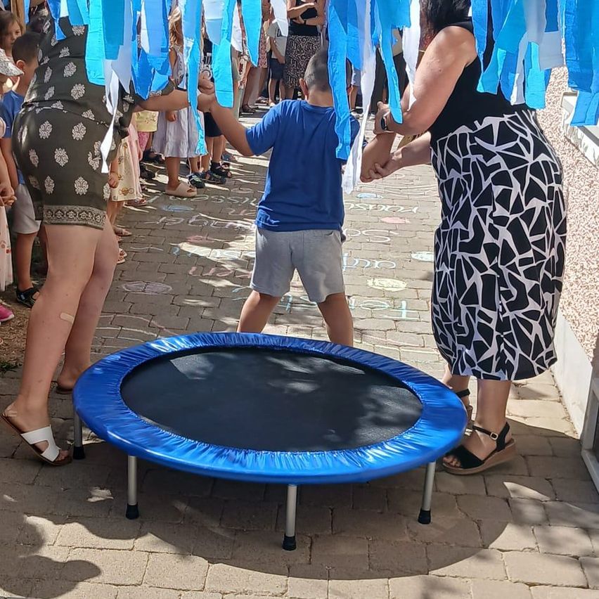 Eine Gruppe von Kindern und Erwachsenen versammelt sich um einen blauen Trampolin auf einem Ziegelboden. Zwei Frauen halten die Hände eines Jungen in einem blauen Shirt. Blaue Bänder hängen über ihnen.