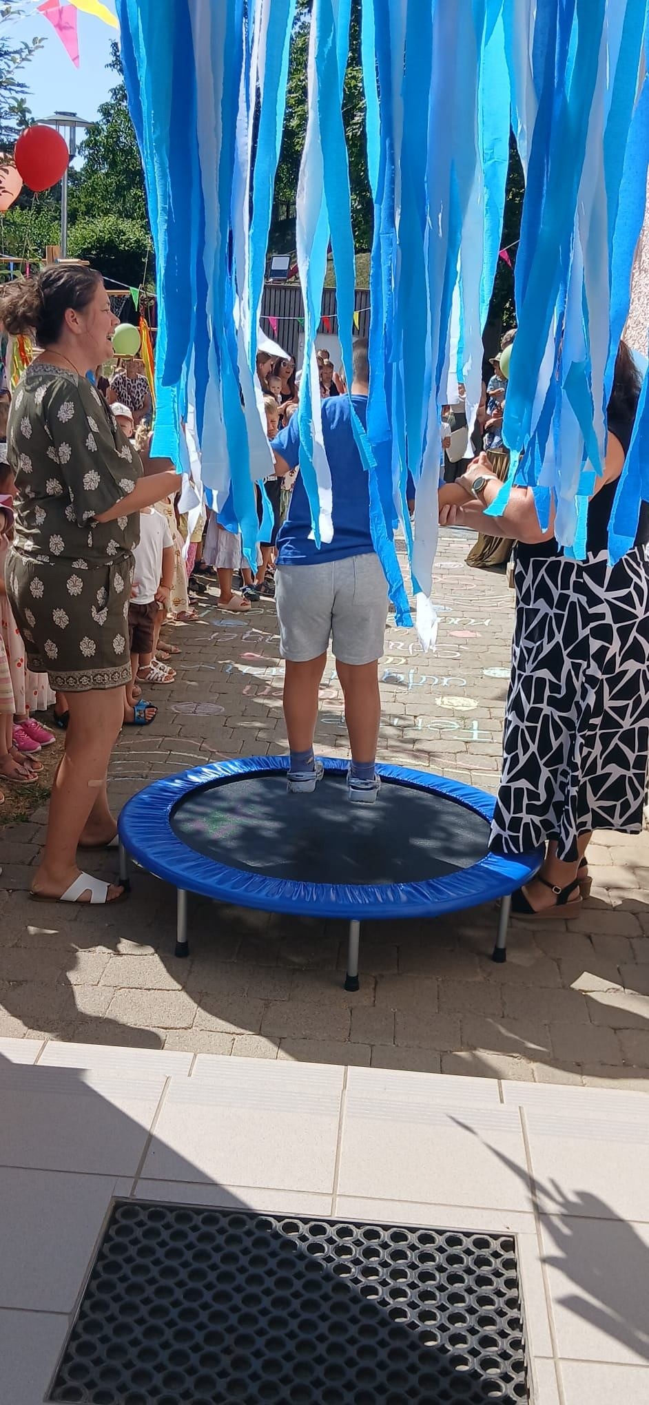 Kinder stehen auf Trampolin unter blauen und weißen Bändern, umgeben von Zuschauern