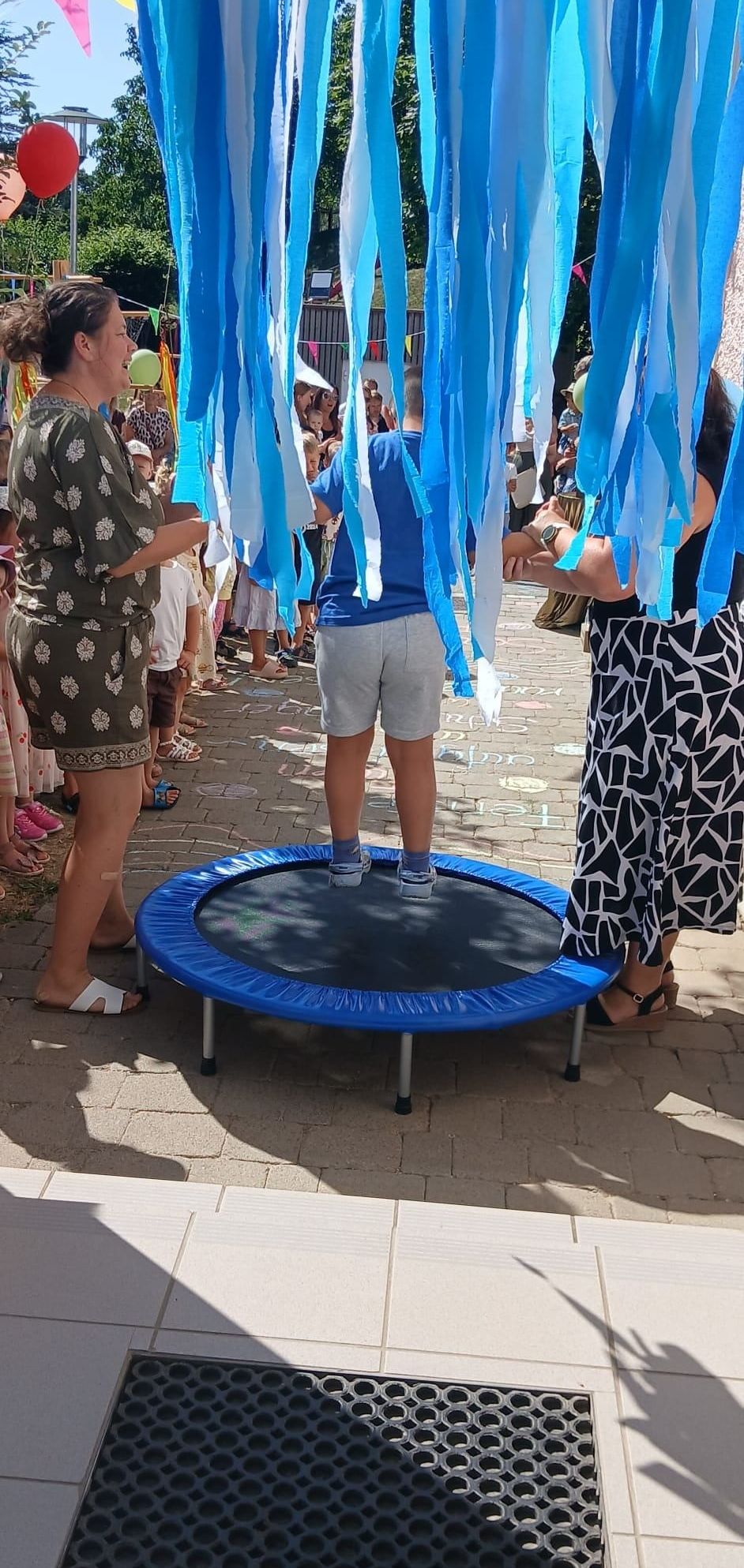 Ein kleiner Junge steht auf einem Trampolin, während Menschen blaue Wimpel über ihm halten. Die Szene ist im Freien mit einem Ziegelboden und einer Menschenmenge im Hintergrund.