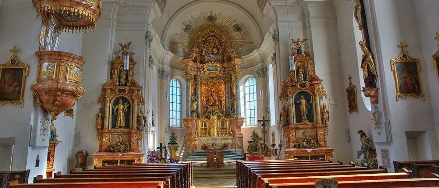 Ein reich verzierter Kircheninnenraum mit goldenen Altären, Statuen und einem Kruzifix. Der Altar ist mit Blumen geschmückt. Bänke sind im Kirchenschiff angeordnet.