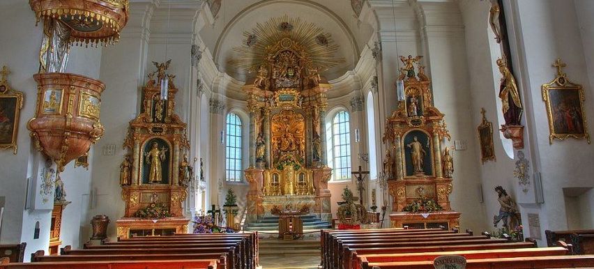 Ein reich verzierter Kircheninnenraum mit goldenen Altären, Statuen und einem Kruzifix. Der Altar ist mit Blumen geschmückt. Bänke sind im Kirchenschiff angeordnet.