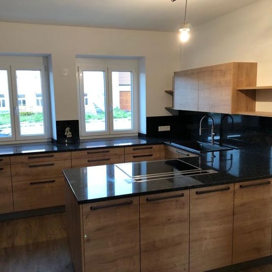 Eine moderne Küche mit Holzschränken, schwarzen Arbeitsplatten und einem Edelstahlspülbecken. Es gibt zwei Fenster mit weißen Rahmen und eine Lampe, die von der Decke hängt.