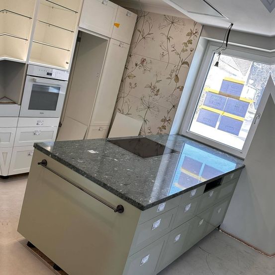 Eine Küche mit einer großen Insel und Granitarbeitsplatte. Sie verfügt über ein modernes Design mit weißen Schränken und Schubladen, einem eingebauten Backofen und einem Fenster mit Blick auf ein Haus.