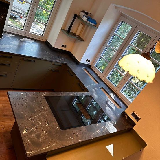 Eine moderne Küche mit dunklen Granitarbeitsplatten und eleganten Schränken. Eine gelbe Lampe hängt von der Decke, und große Fenster lassen viel natürliches Licht herein.