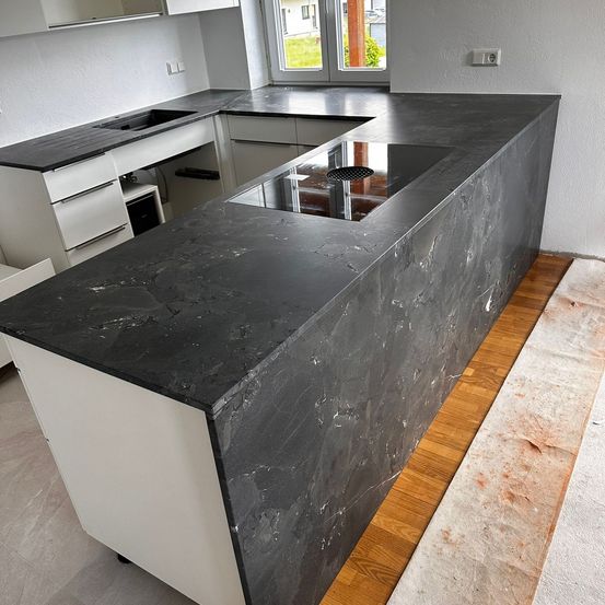 Moderne Küche mit einer großen, eleganten schwarzen Granitarbeitsplatte und einem eingebauten Induktionskochfeld. Weiße Schränke und ein Fenster mit Blick auf die grüne Umgebung.