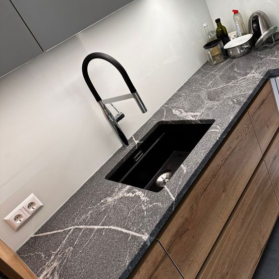 Eine moderne Küche mit grauer Granitarbeitsplatte verfügt über ein elegantes schwarzes Waschbecken mit silbernem Wasserhahn. Die Wand ist weiß und Holzschränke umgeben die Arbeitsplatte.