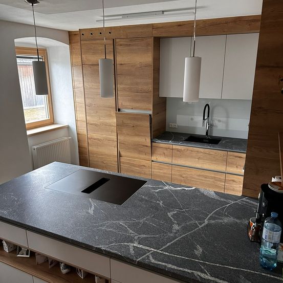 Eine moderne Küche mit Holzschränken, einer Granitarbeitsplatte, einem Waschbecken mit Wasserhahn und an der Decke hängenden Pendelleuchten.