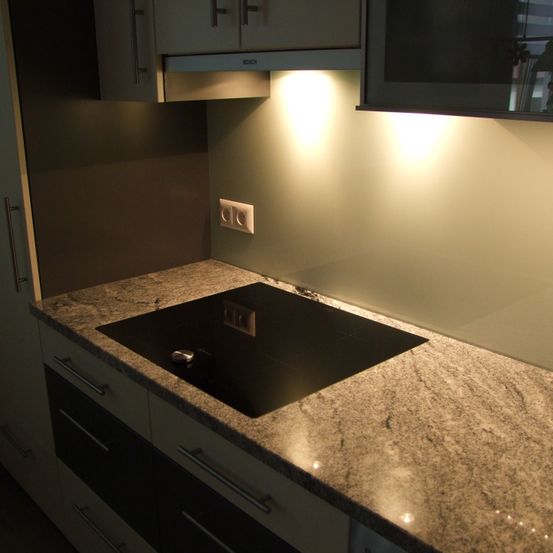 Eine moderne Küche mit Granitarbeitsplatte, schwarzem Induktionskochfeld und weißen Schränken mit silbernen Griffen. Eine weiße Wand wird von einem Licht über dem Herd beleuchtet.