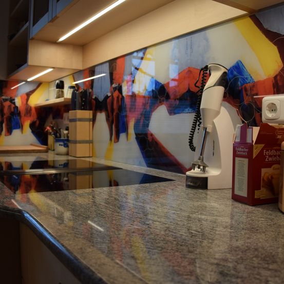 Eine moderne Küche mit Granitarbeitsplatte zeigt ein abstraktes Gemälde an der Wand. Ein weißer Mixer mit Kabel steht auf der Arbeitsplatte neben einer roten Box und einer Steckdose.