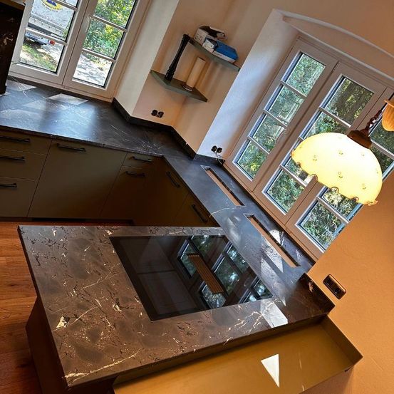 Eine Küche mit Marmorarbeitsplatten, einem Gasherd, einer Lampe und einem Fenster. Die Küche hat einen Holzboden, ein Regal mit Gegenständen und einen Blick auf Bäume draußen.